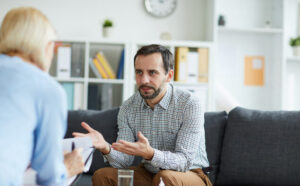 Tense middle aged manager in casualwear explaining his trouble to the Employee Assistance Program psychologist while sitting on couch in front of her